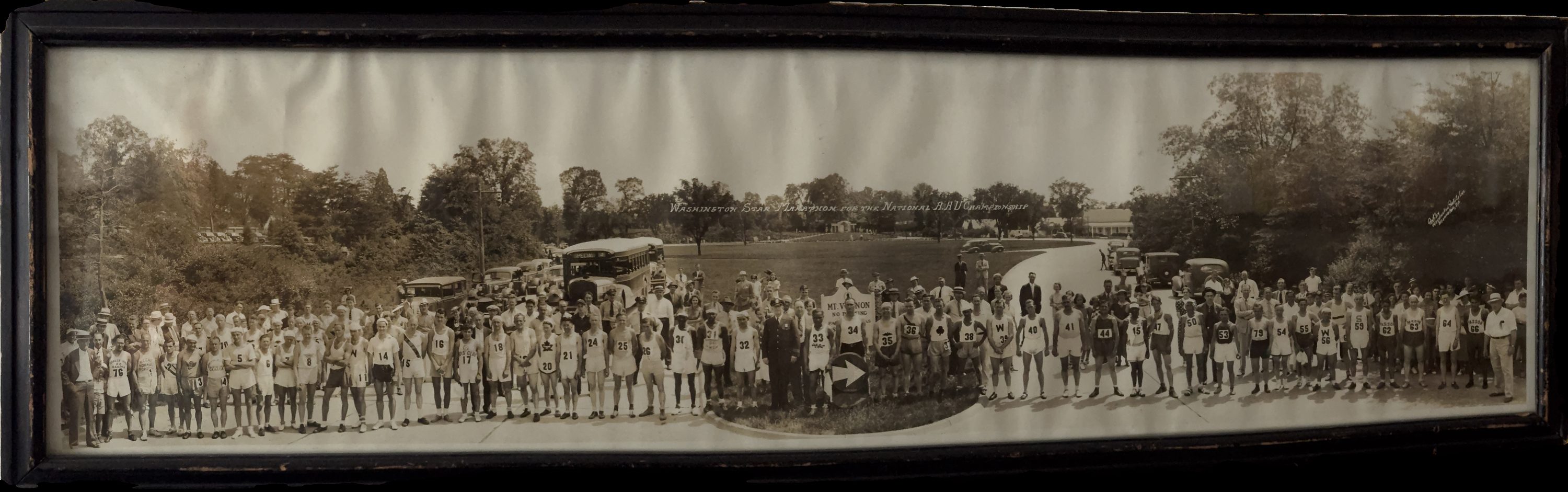 A long panoramic photograph of a marathon field, roughly eighty runners in numbered singlets lined up along a road with buses and cars behind.