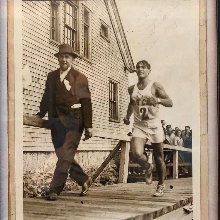 A young dark haired runner in a Boys Club singlet numbered 2 runs along a wooden boardwalk past an official in a hat, with a crowd behind a rail.