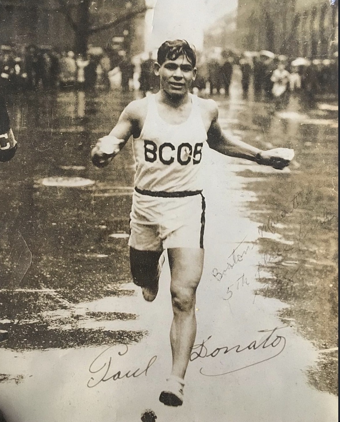 A runner in a BCOB singlet finishing on a wet street lined with spectators under umbrellas. The print is inscribed and signed in the sky.