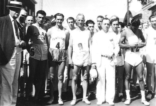A crowd of runners in singlets stands at a street start line among officials in suits and hats.