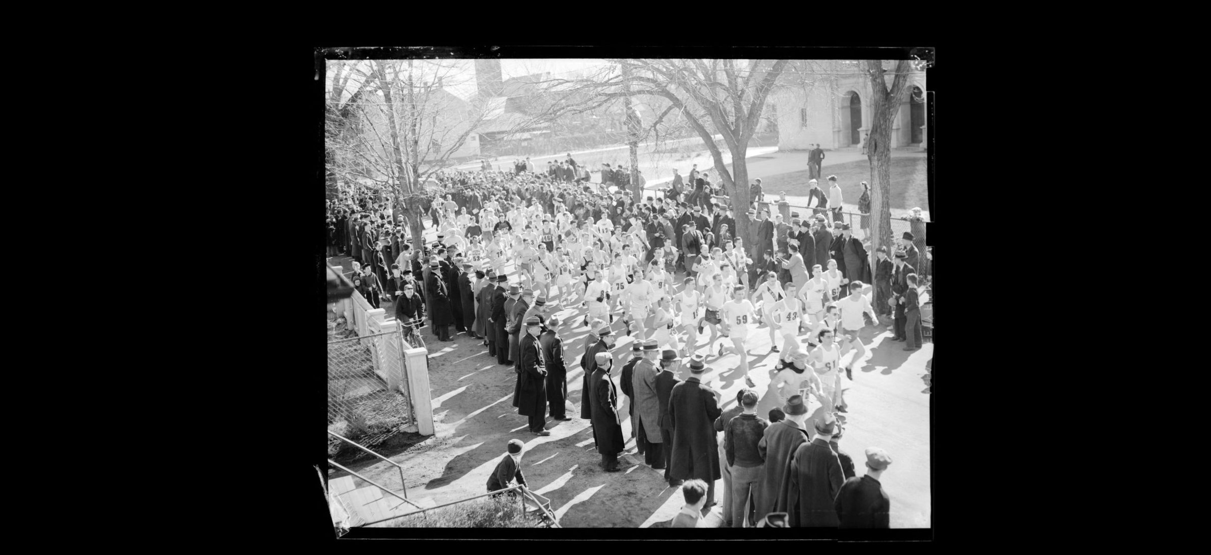 A large field of runners moving away from the start at Hopkinton, spectators lining both sides of the road, bare trees and a civic building behind.