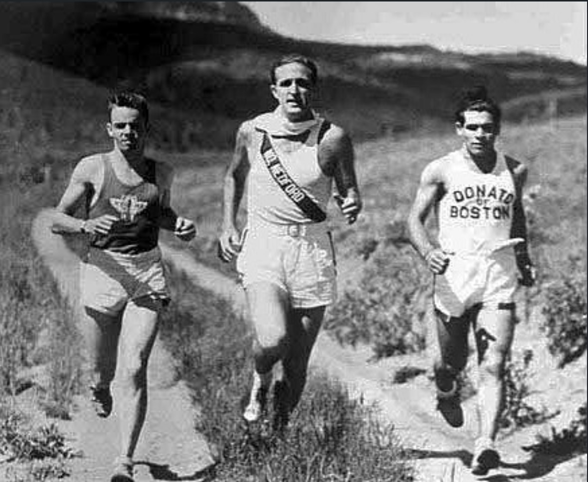 Three runners climbing a dirt road through open scrub. The runner on the right wears a singlet lettered DONATO OF BOSTON.